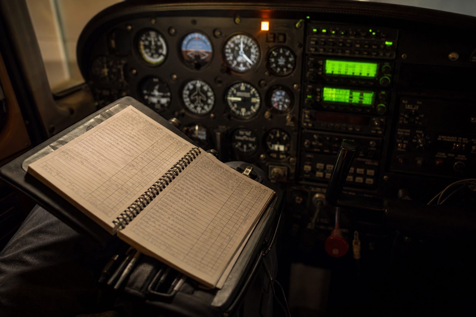 Open pilot logbook on a kneeboard in a dark cockpit lit by green avionics glow and one amber warning light